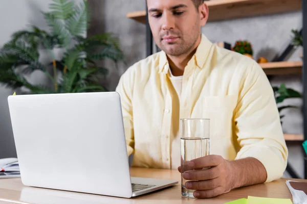 Jovem freelancer trabalhando no laptop perto de vidro de água, fundo borrado — Fotografia de Stock