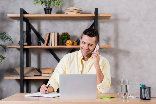 Ein lächelnder Mann telefoniert mit seinem Handy, während er in der Nähe seines Laptops zu Hause arbeitet — Stockfoto