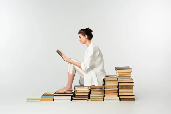 Seitenansicht einer jungen Frau, die auf einem Bücherstapel sitzt, während sie auf Weiß liest — Stockfoto