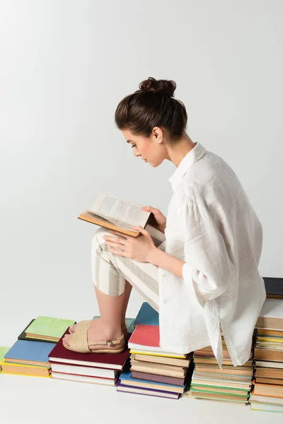 Young brunette woman reading while sitting on stack of books isolated on white — Stock Photo