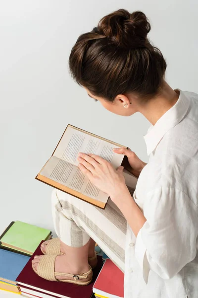 High angle view of young woman reading while sitting on stack of books isolated on white — Stock Photo