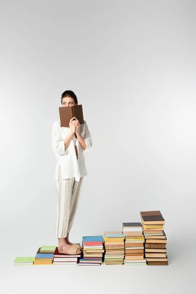 Full length of young woman in glasses standing on pile of books while covering face on white — Stock Photo