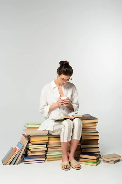 Full length of young woman in glasses sitting on a pile of books while reading and holding cup on white — стоковое фото