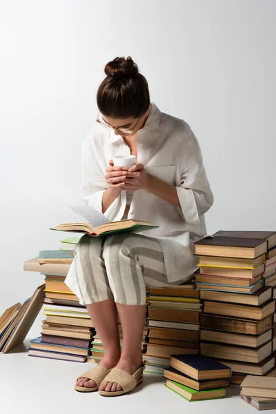 Full length of young brunette woman in glasses sitting on pile of books while reading and holding cup on white — Stock Photo
