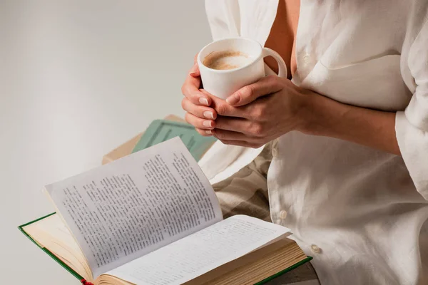 Cropped view of young woman reading book and holding cup of coffee isolated on white — Stock Photo