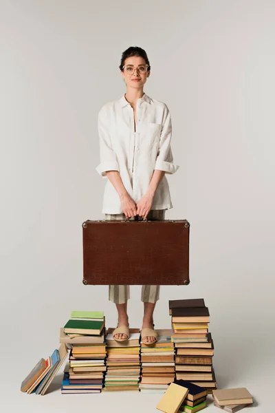 Longitud completa de la joven complacida en gafas que sostienen la maleta mientras está de pie sobre una pila de libros aislados en blanco - foto de stock