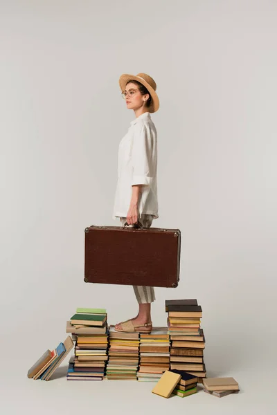 Full length of young woman in straw hat and glasses holding suitcase while standing on pile of books isolated on white — Stock Photo