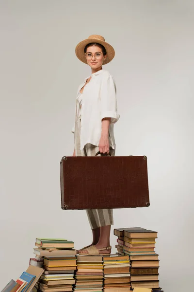 Pleine longueur de femme heureuse en chapeau de paille et lunettes tenant bagages tout en se tenant debout sur une pile de livres isolés sur blanc — Photo de stock
