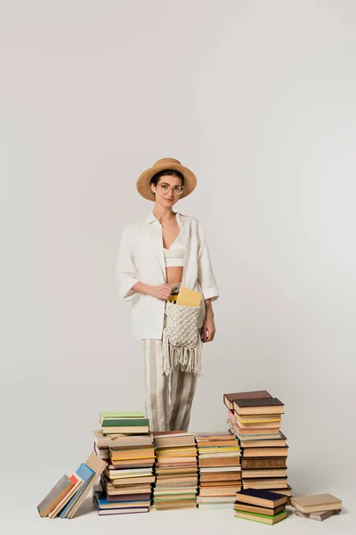 Giovane donna in cappello da sole in piedi vicino mucchio di libri isolati su bianco — Foto stock