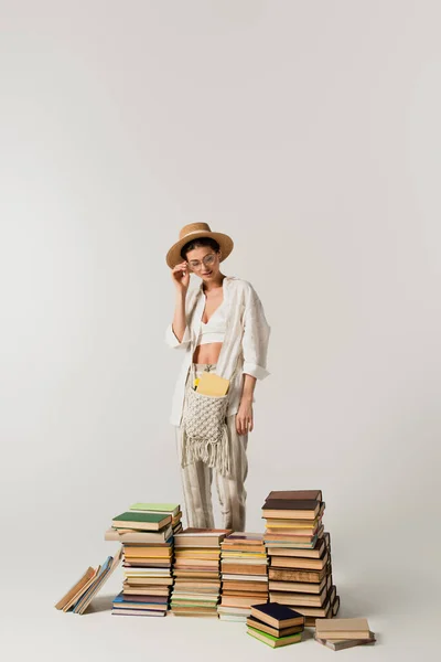 Bonita mujer joven en sombrero de sol de pie cerca de pila de libros aislados en blanco - foto de stock