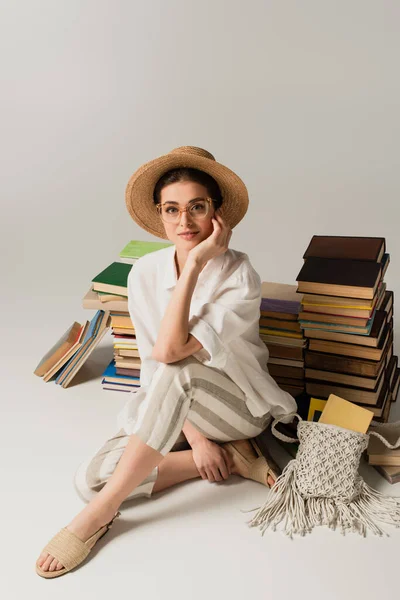 Full length of pleased young woman in sun hat and glasses sitting near pile of books on white — Stock Photo