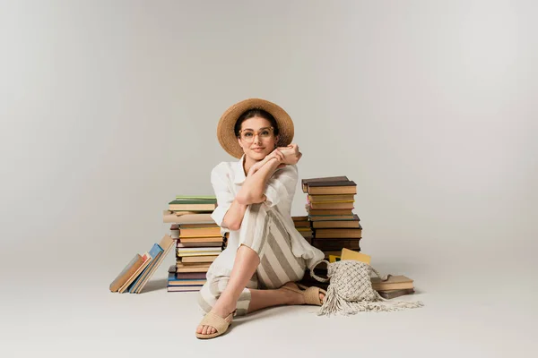 В полный рост счастливая молодая женщина в солнцезащитной шляпе и очках, сидящая возле стопки книг на белом — стоковое фото