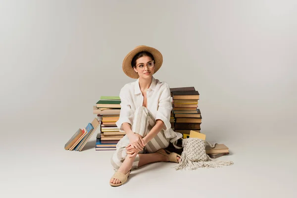 Full length of happy young woman in straw hat and glasses sitting near stack of books on white — Stock Photo