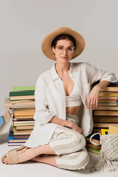 Pleased woman in straw hat and glasses sitting near stack of books on white — Stock Photo