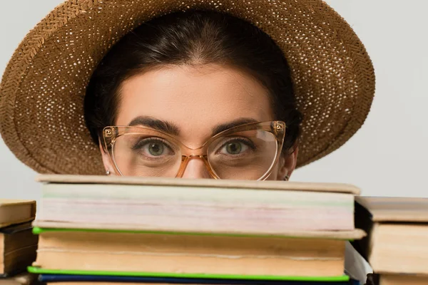 Junge Frau mit Sonnenhut und Brille blickt durch einen Stapel Bücher isoliert auf weiß in die Kamera — Stockfoto