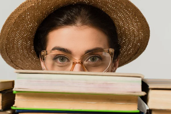 Young woman in sun hat and glasses looking at camera through pile of books isolated on white — Stock Photo