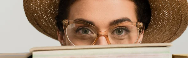 Giovane donna in cappello da sole e occhiali guardando la macchina fotografica attraverso mucchio di libri isolati su bianco, banner — Foto stock