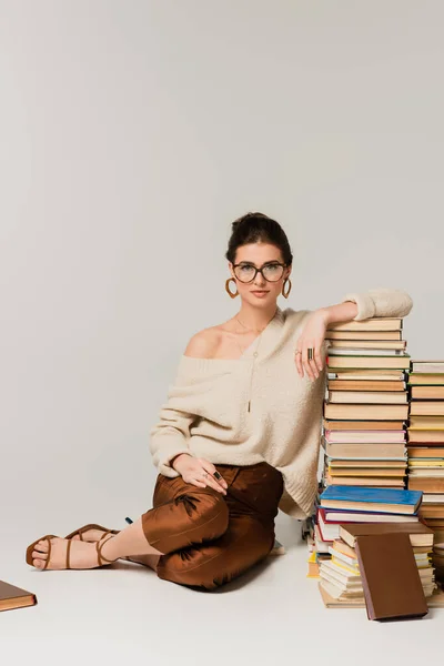 Full length of young woman in glasses and sweater leaning on pile of books on white — Stock Photo