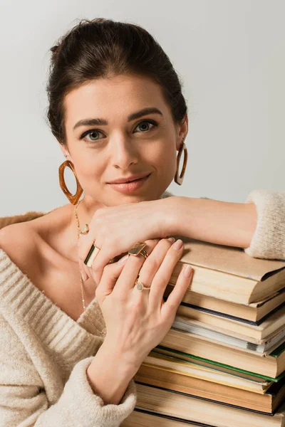 Улыбающаяся молодая женщина в серьгах, опирающаяся на стопку книг, изолированных на белом — стоковое фото