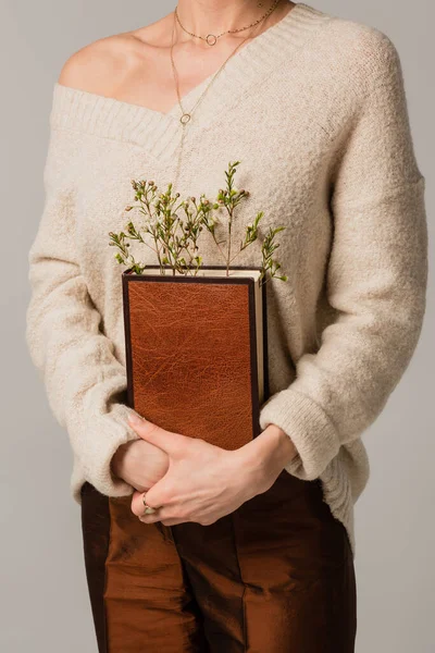 Vista recortada de la joven mujer sosteniendo libro con flores silvestres aisladas en gris - foto de stock