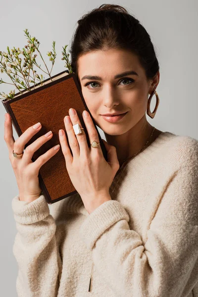 Smiling young woman holding book with wildflowers isolated on grey — Stock Photo