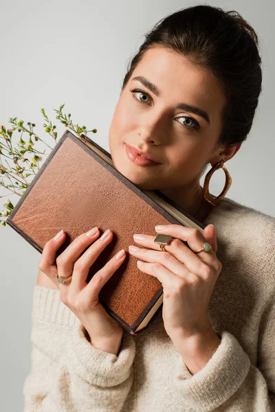 Радостная молодая женщина держит книгу с полевыми цветами, изолированными на сером — стоковое фото