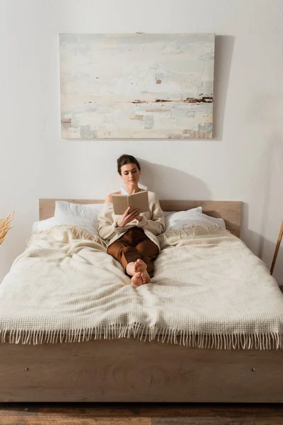 Barfüßige junge Frau liest Buch, während sie zu Hause im Bett liegt — Stockfoto