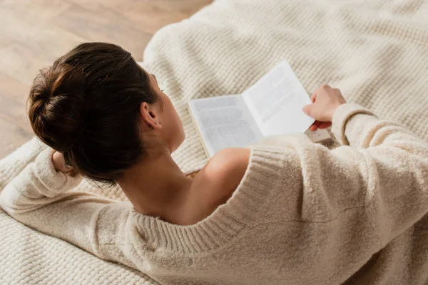 Back view of young brunette woman reading book while resting on bed at home — Stock Photo