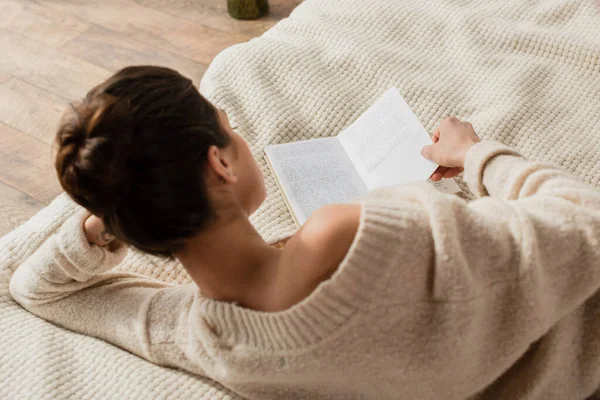 Вид ззаду молода брюнетка в светрі читає книгу, відпочиваючи на ліжку вдома — стокове фото