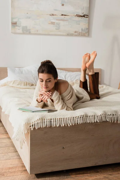 Barfüßige junge Frau liest Buch, während sie sich im Bett ausruht — Stockfoto
