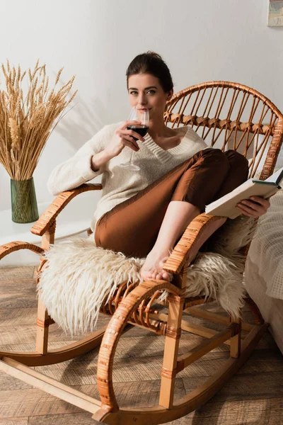 Junge Frau sitzt im Weidenschaukelstuhl mit Buch und trinkt Rotwein — Stockfoto
