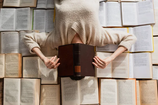 Top view of tattooed woman obscuring face while lying on pile of open books — Stock Photo