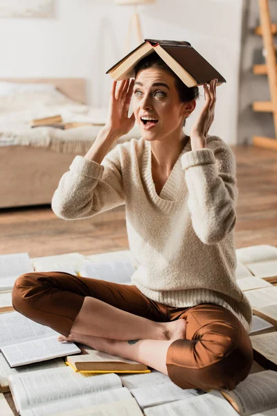 Amazed woman with book on head sitting on pile of open pages — Stock Photo