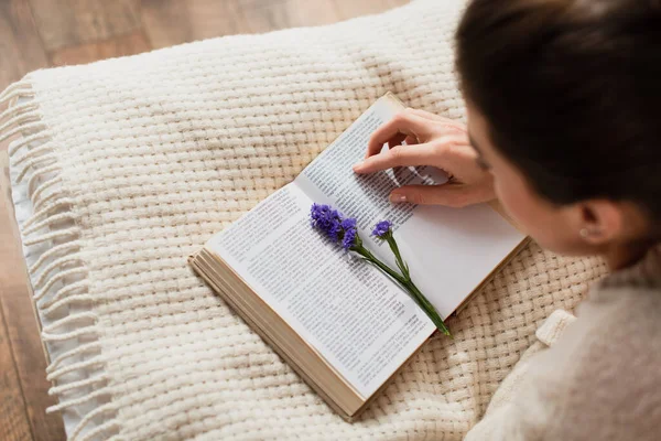 Високий кут зору розмита молода жінка читає книгу з фіолетовою квіткою під час відпочинку на ліжку вдома — стокове фото