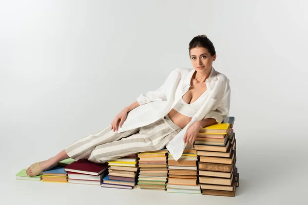 Full length of brunette young woman in sandals lying on pile of books on white — Stock Photo
