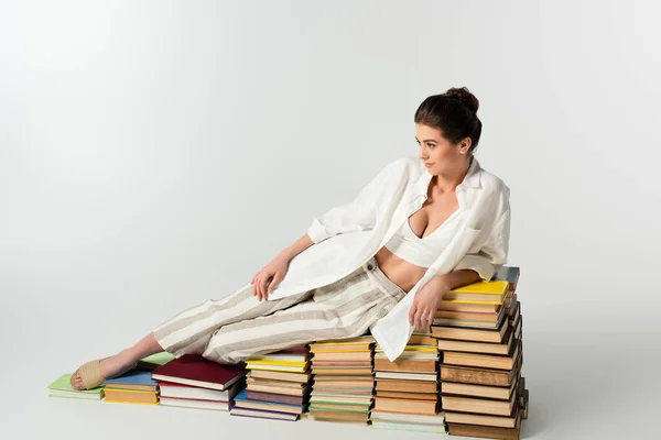 Full length of young brunette woman in sandals lying on pile of books on white — Stock Photo