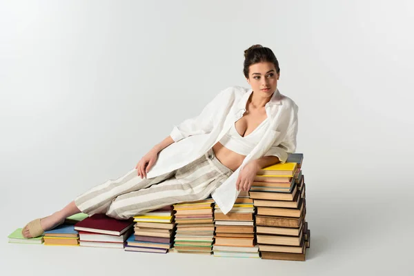 Full length of young woman in sandals lying on a pile of books on white — стоковое фото