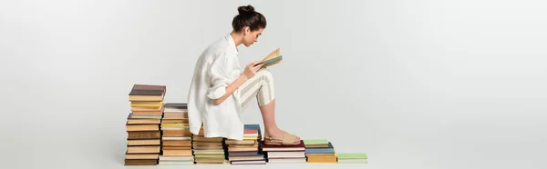 Side view of young woman in sandals reading while sitting on pile of books on white, banner — Stock Photo