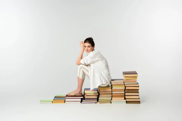 Longitud completa de la joven sonriente en sandalias sentadas en un montón de libros en blanco - foto de stock