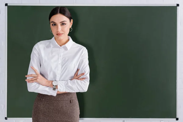 Insegnante con le braccia incrociate guardando la macchina fotografica vicino alla lavagna in classe — Foto stock