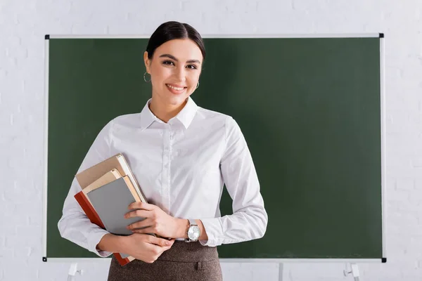 Teacher with books smiling at camera in classroom — Stock Photo