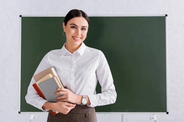 Professeur avec des livres souriant à la caméra dans la salle de classe — Photo de stock