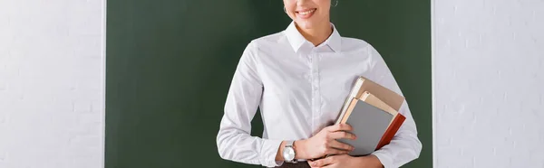 Vista recortada del profesor sonriente con libros de pie cerca de pizarra, pancarta - foto de stock