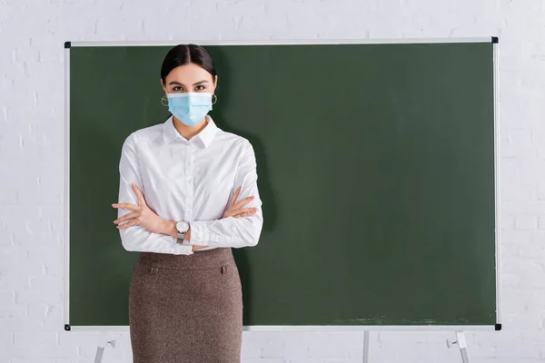 Учитель в медицинской маске стоит со скрещенными руками возле доски — стоковое фото