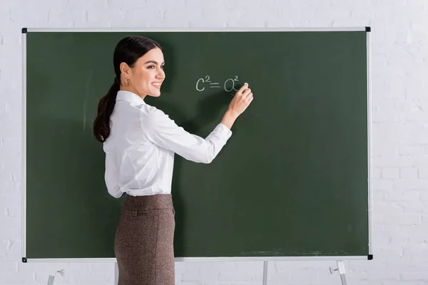 Cheerful teacher with chalk writing mathematic equation on chalkboard — Stock Photo