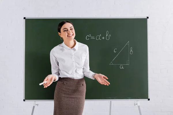 Smiling teacher with chalk standing near chalkboard during lesson — Stock Photo