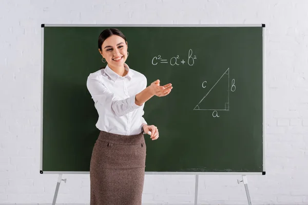 Smiling teacher pointing with hand near chalkboard during mathematic lesson — Stock Photo