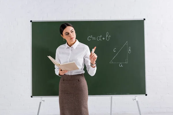 Profesor con libro y tiza señalando con el dedo durante la lección en el aula - foto de stock