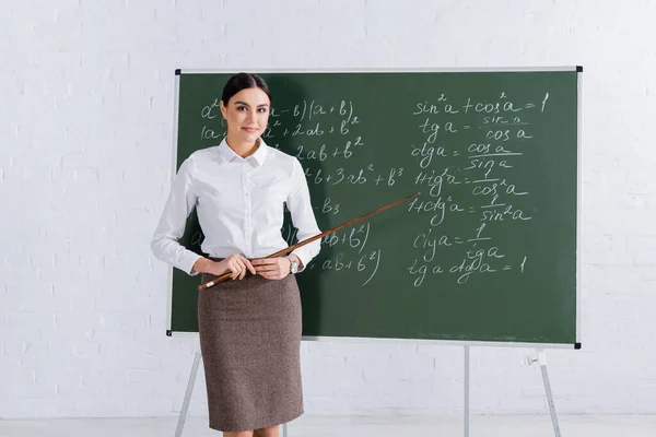 Profesor con puntero sonriendo a la cámara cerca de pizarra durante la lección matemática - foto de stock