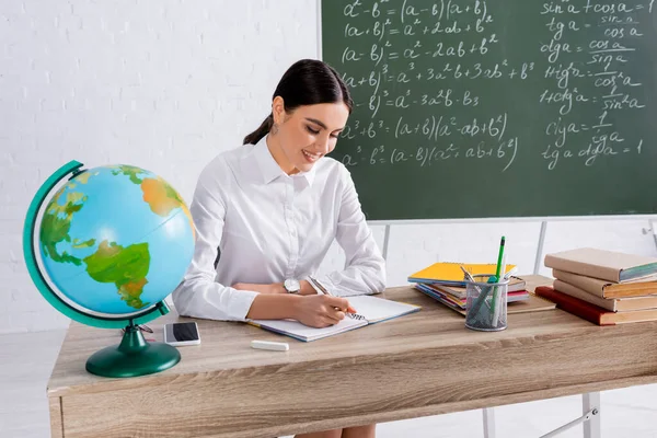 Joven profesor escribiendo en un cuaderno cerca del mundo, teléfonos inteligentes y libros - foto de stock